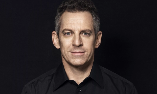 A man with short gray hair and a slight smile, wearing a black shirt, against a dark background.