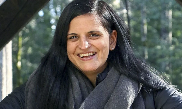 A woman with long black hair, wearing a dark jacket and gray scarf, smiles outdoors surrounded by trees.