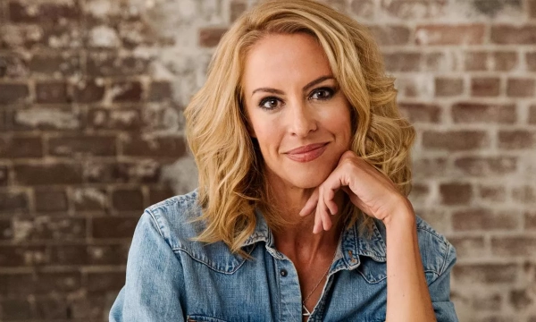 A woman with blonde, wavy hair smiles while resting her chin on her hand, wearing a denim jacket against a brick wall backdrop.
