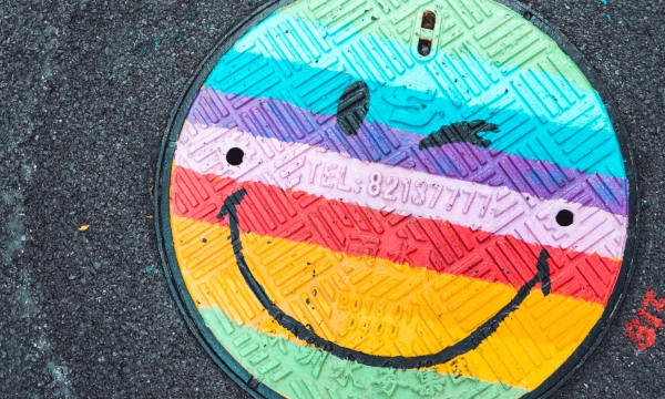 A colorful manhole cover featuring a smiling face design, painted in vibrant rainbow stripes, set against a dark asphalt background.