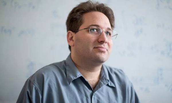 A man with glasses and a gray shirt, sitting against a whiteboard filled with mathematical equations.
