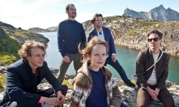 Group of five individuals in casual clothing posing by a rocky shoreline with water and mountains in the background, suggesting a scenic outdoor setting.