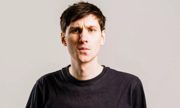 A young man with short brown hair, wearing a plain black t-shirt, stands against a light gray background, looking seriously at the camera.