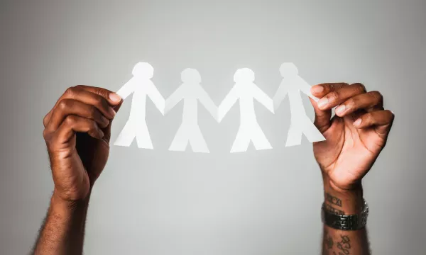 A person's hands hold a chain of four paper cutout figures, symbolizing unity and connection, against a plain gray background.