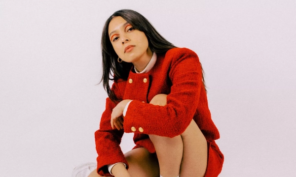 A young woman poses in a red outfit, crouching with one knee up, wearing white sneakers and showing a confident expression against a plain white background.