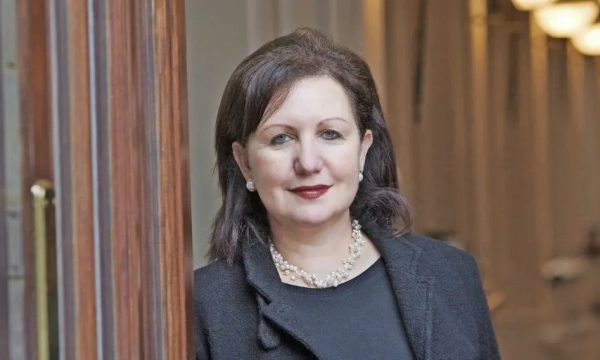 Confident woman with dark hair, wearing pearl earrings and a black coat, posed in an elegant indoor setting with warm lighting.