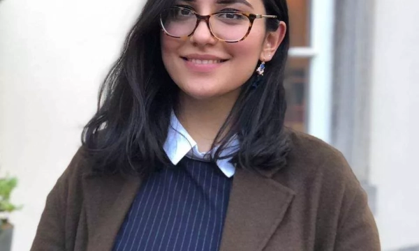 A smiling young woman with glasses and long dark hair, wearing a brown coat over a pinstripe shirt, stands outdoors in a well-lit setting.