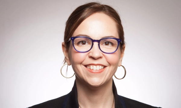 A smiling woman with glasses and hoop earrings, wearing a black blazer, poses against a neutral background.