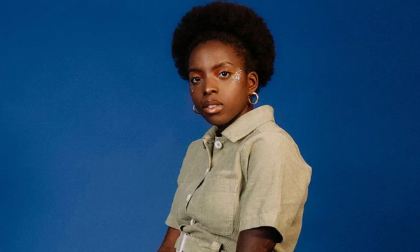 A young person with an afro hairstyle poses against a blue background, wearing a light green shirt, showcasing a confident expression.