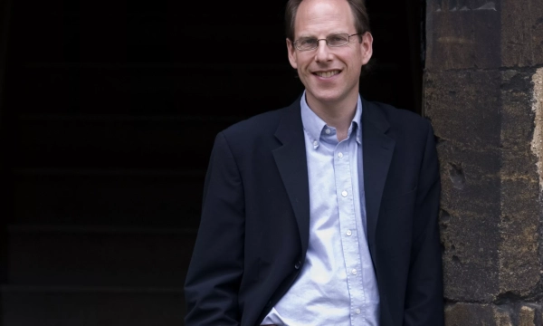 A man wearing a blazer and glasses stands in front of a stone archway, smiling gently as he leans against the wall.