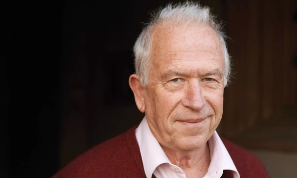 Elderly man with gray hair, wearing a maroon sweater over a pink shirt, smiling and standing against a dark background.