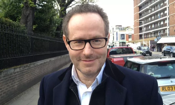 A man wearing glasses and a dark coat smiles at the camera while standing on a city street with vehicles and buildings in the background.