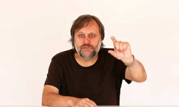 A man with a beard and long hair gestures while seated at a table, against a plain background, conveying a thoughtful expression.