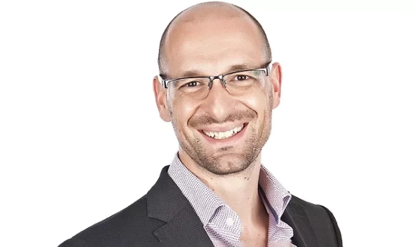 A smiling man with glasses, wearing a dark suit jacket over a light-colored shirt, poses against a plain white background.