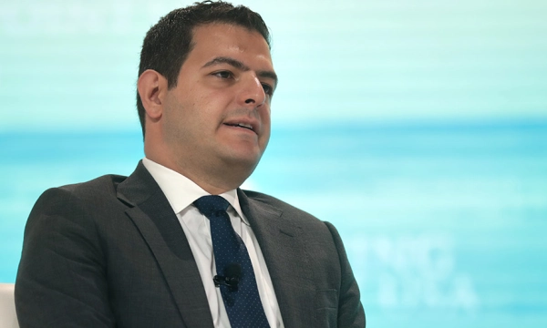 A well-dressed man in a gray suit and blue tie sits on stage during a panel discussion, with a light blue background and microphone visible.