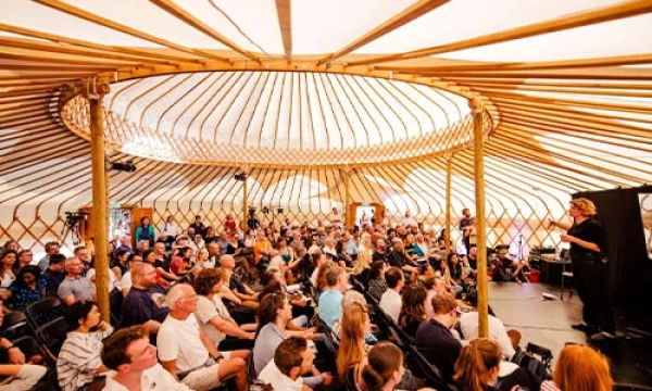 A large audience gathers in a yurt for a presentation, featuring rows of chairs and a speaker on stage discussing various topics.