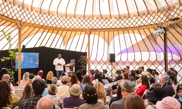 A speaker presents to a large audience inside a spacious, wooden yurt, surrounded by natural light filtering through the structure's unique design.