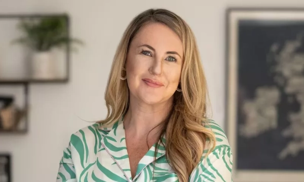 A smiling woman with long, wavy hair, wearing a green and white patterned top, stands confidently in a well-lit room with plants and artwork in the background.