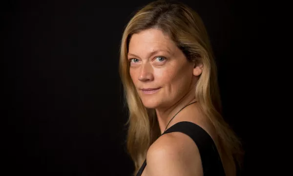 A woman with long, blonde hair poses against a black background, looking over her shoulder with a subtle smile.