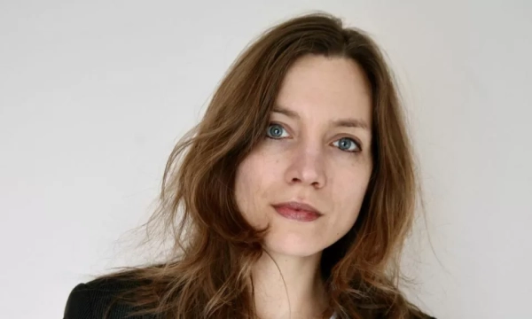 A woman with long, wavy hair and blue eyes poses against a plain background, wearing a black blazer.