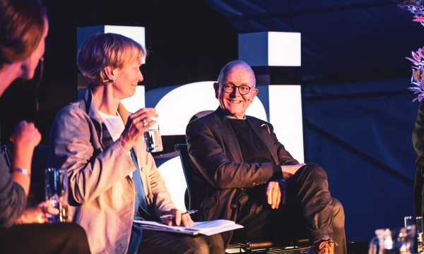 A panel discussion featuring two individuals, one holding a glass of water, while an older man smiles warmly, seated under ambient lighting.
