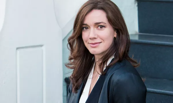 Woman with long brown hair, wearing a black jacket over a white top, sitting on dark stairs, looking confidently at the camera.