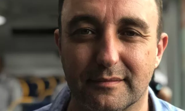 A man with short dark hair and a slight stubble is looking directly at the camera while seated, wearing a light blue shirt.