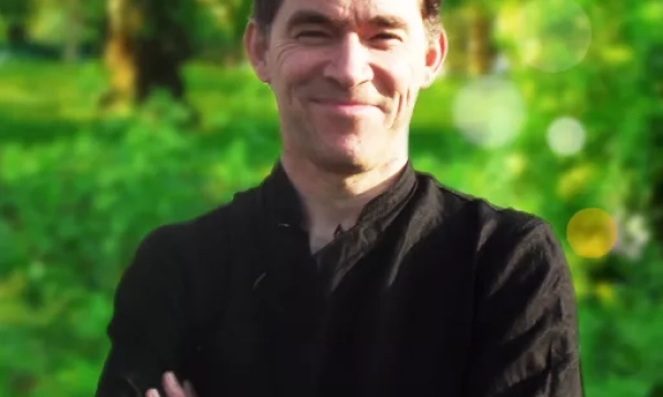 A smiling man in a black shirt stands outdoors, surrounded by greenery and soft light, suggesting a peaceful, natural setting.