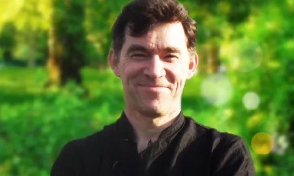 A smiling man with dark hair, dressed in a black shirt, stands outdoors against a green, natural background.