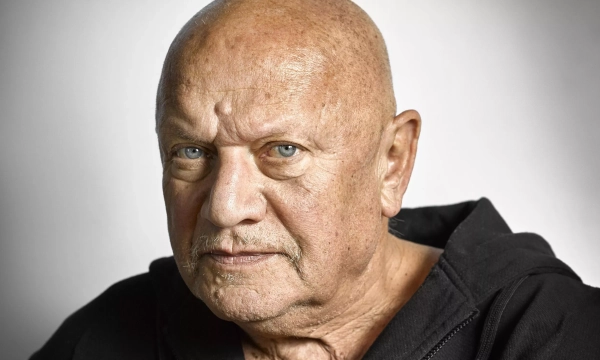 Close-up portrait of a bald man with blue eyes and a serious expression, wearing a black hoodie against a gradient background.