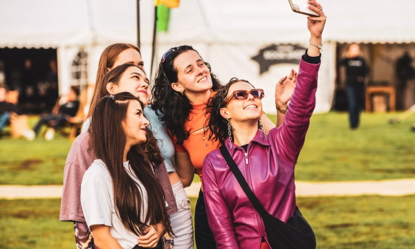 Five young women take a selfie together outdoors at a festival, smiling and wearing colorful outfits.