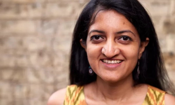 A smiling woman with long black hair, wearing an ethnic patterned top and earrings, stands in front of a textured brick background.