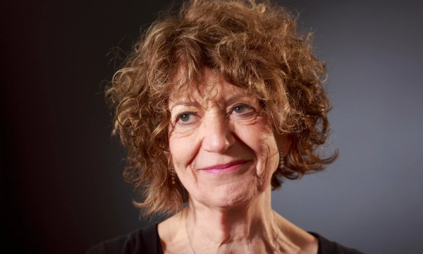 A middle-aged woman with curly hair smiles gently against a soft-focus dark background, exuding warmth and wisdom.