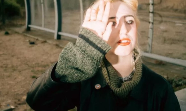 A young woman with blonde hair and bold red lips raises her hand to shield her eyes from the sunlight, seated in a desolate, overgrown area with a modern structure in the background.