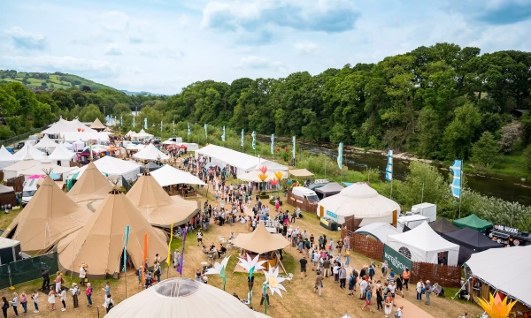Aerial view of a vibrant festival scene, featuring large tents, food stalls, and crowds of people enjoying the outdoors by a river surrounded by greenery.