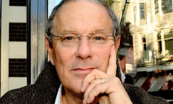 A thoughtful man with glasses and a hand on his chin, seated outside a café with urban scenery in the background.