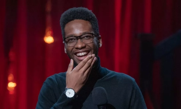 A smiling man in a black turtleneck and glasses holds his hand to his chin while standing on a stage with red curtains in the background.