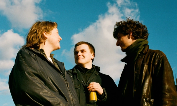 Three people stand outdoors against a blue sky with scattered clouds, engaged in conversation and smiling, with one holding a can.