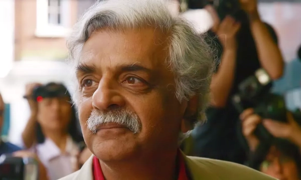 A middle-aged man with silver hair and a mustache, wearing a light-colored coat, surrounded by photographers and onlookers in an urban setting.
