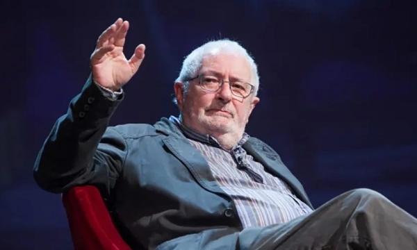 An older man with glasses and white hair is seated, gesturing with one hand while wearing a jacket and striped shirt.