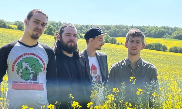 A group of four men stands in a yellow flower field on a sunny day, with green hills in the background.