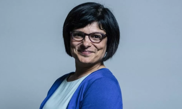 A woman with short black hair and glasses smiles while wearing a blue cardigan against a neutral background.