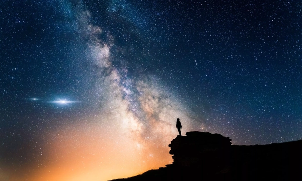 A silhouetted figure stands on a rock, gazing at the vibrant Milky Way and star-filled sky during twilight.