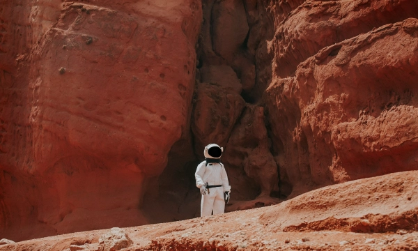 An astronaut in a white spacesuit stands on a rocky, reddish landscape, surrounded by towering cliffs and dramatic rock formations.