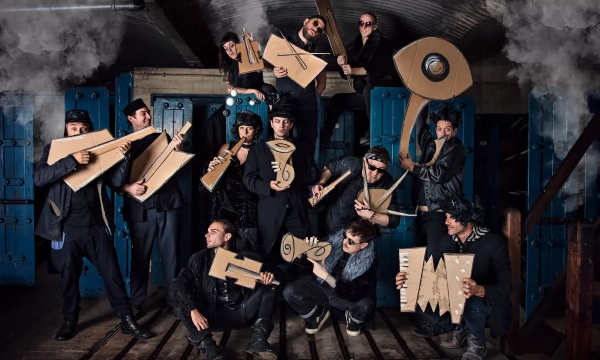 A group of musicians poses with oversized cardboard instruments in a dimly lit industrial setting, creating a playful and artistic atmosphere.