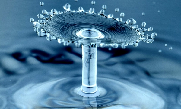 A close-up of a water droplet creating a splash, forming a distinctive mushroom-like shape with numerous tiny droplets surrounding it in a blue-toned environment.