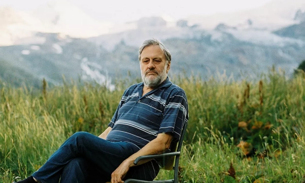 A contemplative man sitting in a chair outdoors, surrounded by tall grass and mountains in the background.