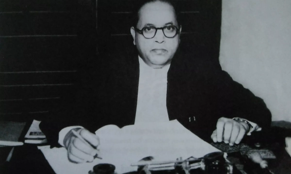A black-and-white image of a man with glasses sitting at a desk, examining documents, possibly a prominent historical figure or politician.