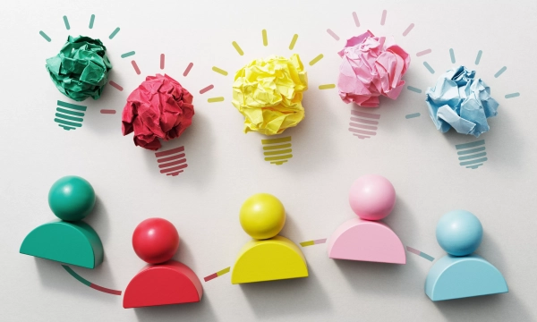Colorful paper crumples shaped like light bulbs above stylized figures, symbolizing ideas, teamwork, and creativity in a collaborative environment.