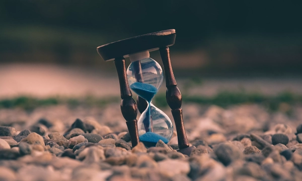 An hourglass with blue sand sits atop a bed of pebbles, symbolizing the passage of time in a serene outdoor setting.